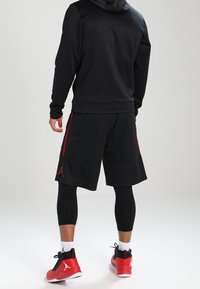 Sudadera deportiva negra con un diseño ajustado, combinada con pantalones cortos negros con acentos rojos, mallas de compresión negras y zapatos rojos.