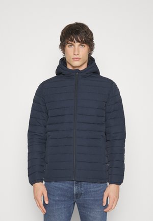 Giubbotto puffer di colore blu navy con cappuccio, caratterizzato da cuciture a righe orizzontali, una zip frontale e polsini elastici. Indossato sopra un paio di jeans.