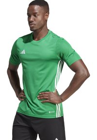 Maglietta sportiva verde realizzata in tessuto leggero, con maniche corte, collo rotondo, tre righe bianche sui lati e logo Adidas.