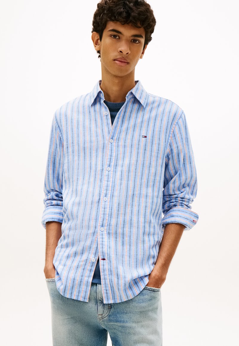 Νεαρός άνδρας που φοράει ανοιχτό μπλε πουκάμισο με ρίγες πάνω από σκούρο μπλε μπλουζάκι και ανοιχτόχρωμα τζιν, με τα χέρια στις τσέπες, ουδέτερο φόντο.