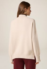 Beżowy oversize sweter z wysokim dekoltem, prążkowanymi mankietami i dolnym wykończeniem. Miękka tekstura, luźny krój, w zestawieniu z burgundowymi spodniami.