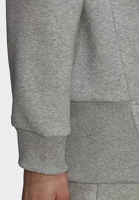 Gros plan sur la main et le bas du bras d'une personne portant un sweat gris clair avec des poignets et une ceinture c ôtelés.