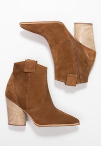 Bottines en daim marron avec un bout pointu, un talon bloc strié, des détails festonnés et une fermeture à glissière sur le côté.