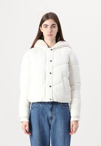 White puffer jacket with a soft, fur-lined hood, featuring a snap-button closure and diagonal stitching. Worn with blue denim jeans.