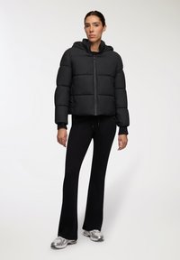 Kobieta stoi w czarnej kurtce puchowej z kapturem, czarnych spódnicach w kształcie dzwonu i srebrnych butach sportowych na białym tle.