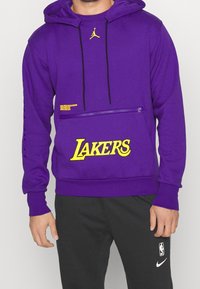 Sudadera con capucha morada hecha de una tela con acabado texturizado. Presenta un logotipo amarillo de "LAKERS" y detalles con bolsillo con cremallera.