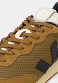 Sneaker marrone con tomaia in rete testurizzata, accenti crema e dettagli blu scuro. Presenta lacci e suola in gomma con logo V.