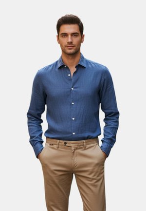 Uomo che indossa una camicia blu a maniche lunghe abbottonata, infilata in pantaloni beige, con le mani in tasca, in piedi contro uno sfondo neutro.