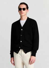 Cardigan noir en tricot avec un col en V, comprenant six boutons, des poignets et un ourlet côtelés ; porté sur une chemise blanche et associé à un pantalon crème.