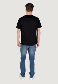 T-shirt nera a maniche corte in cotone vista dal retro, abbinata a jeans in denim azzurro chiaro e sneakers bianche, su uno sfondo neutro.