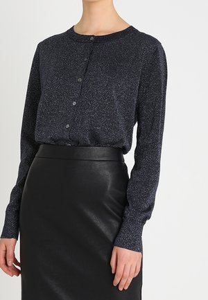Kvinde iført en mørk glitrende langærmet bluse med knapper, stukket ned i et højtaljet sort lædernederdel.