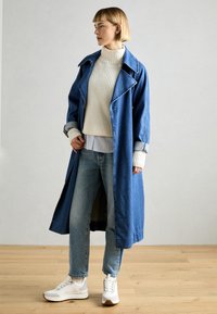 Abrigo largo de mezclilla azul con cuello muesca, combinado con un suéter blanco de canalé y jeans azul claros. Zapatillas blancas completan el atuendo.