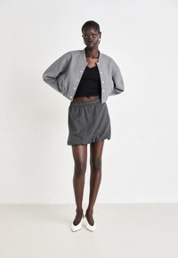 Veste bomber grise, courte, avec des boutons-pression, top en dentelle noire, mini-jupe rayée et chaussures à talons blancs. Design simple et moderne.
