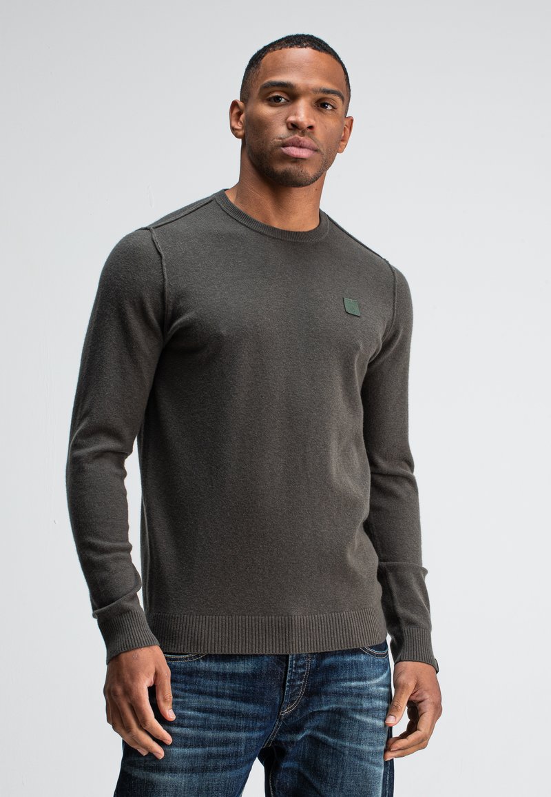 Dunkelgrauer Pullover mit langen Ärmeln und gerippten Bündchen, mit einem kleinen, quadratischen Logo auf der Brust, kombiniert mit blauen Jeans.