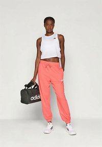 Athletin in einem weißen Crop-Top und korallenfarbenen Jogginghosen, die eine schwarze Adidas-Duffle-Tasche hält. Die Jogginghosen haben einen elastischen Bund und ein Logo.