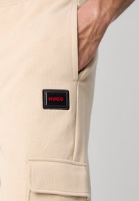Beige cargobyxor i mjuk bomullsblandning, med sidoficka och en svart lapp med "HUGO" i rött. Rena linjer, avslappnad design.