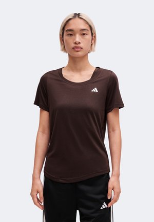Stående person med kort blondt hår iført en mørkebrun Adidas t-shirt og sorte Adidas bukser mod en ensfarvet lys baggrund.