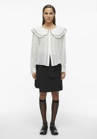 Blusa blanca y ligera con un gran cuello arrugado y ribete negro, combinada con una falda negra que tiene un bolsillo delantero y calcetas hasta la rodilla.