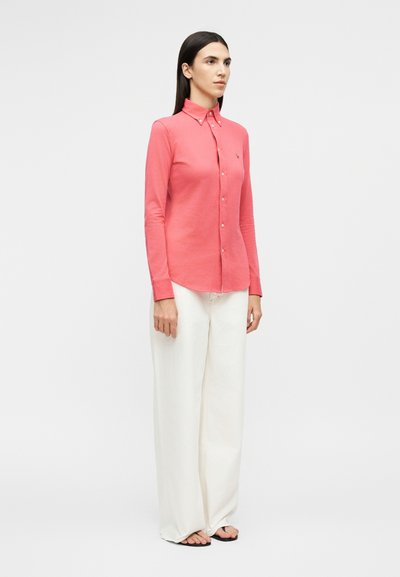 Nő áll, korallrózsaszín gombos hosszú ujjú inget, bő szárú fehér nadrágot és fekete papucsot visel, egyszerű háttér előtt.