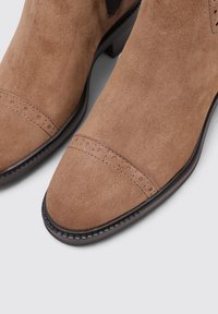 Botas de ante marrón hasta el tobillo con punta redonda, que presentan un patrón de brogue cerca de la puntera y una suela oscura texturizada.