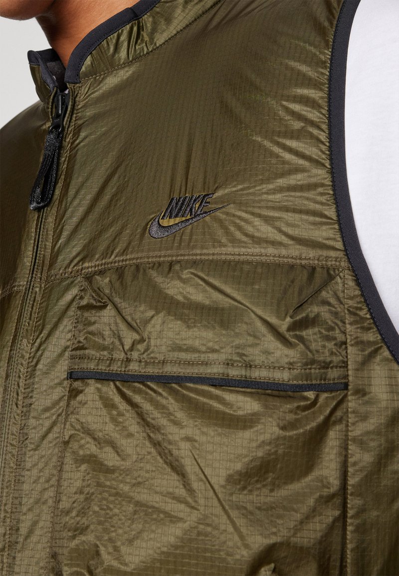 Gros plan sur une veste sans manches Nike vert olive avec une fermeture éclair noire, une poche avant et un logo sur la poitrine portée sur une chemise blanche.