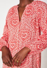 De handen van een persoon die een rode en witte geblokte blouse met lange mouwen en elastische manchetten draagt, met een gouden ring met een rechthoekige steen.