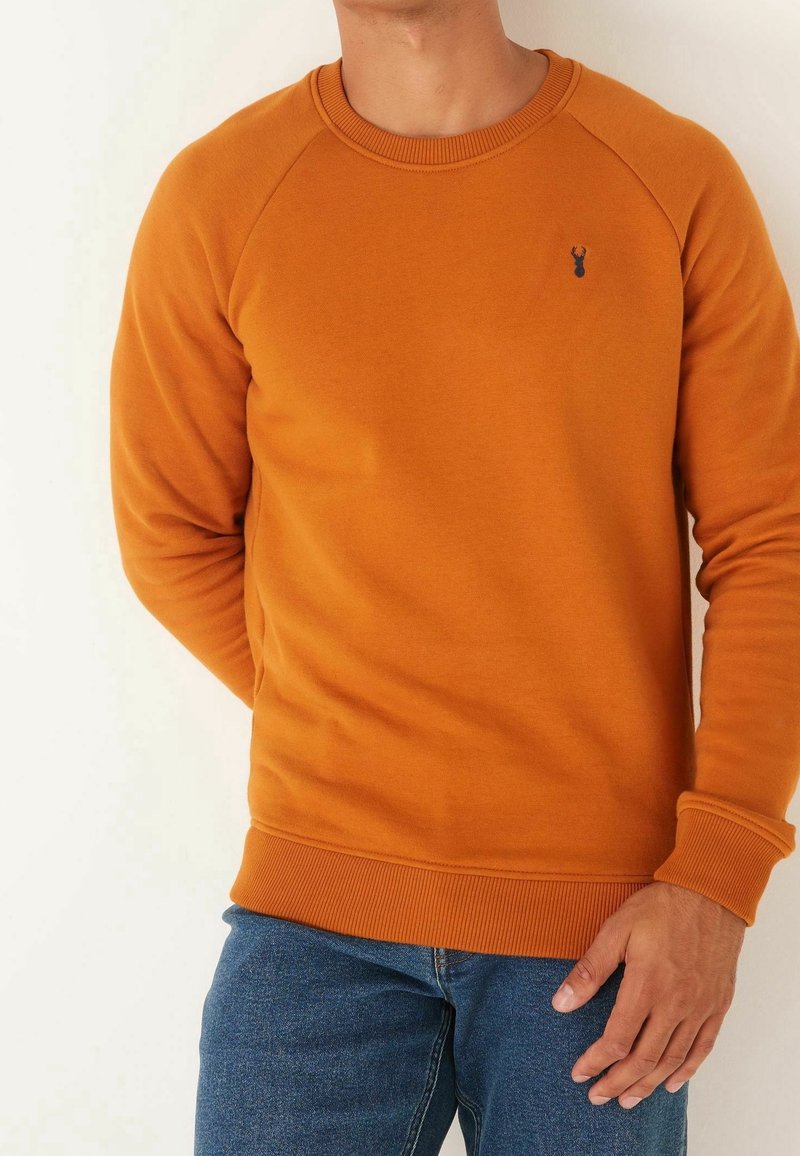Homme portant un pull à manches longues orange moutarde avec poignets et ourlet côtelés, associé à un jean bleu, sur un fond uni blanc cassé.