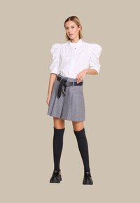 Hvit skjorte med puff-sleeves, grå plisserte skjørt med svart belte, knelange mørke sokker og svarte sko. Materialet ser glatt og strukturert ut.