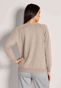 Frau mit langen dunklen Haaren, die einen beigen Pullover und hellgraue Hose trägt, mit einer Hand auf der Hüfte stehend, von hinten gesehen.