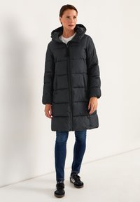 Svart puffjacka med huva, långa ärmar, dragkedja fram och horisontell quiltning. Matchad med blå jeans och svarta sneakers.