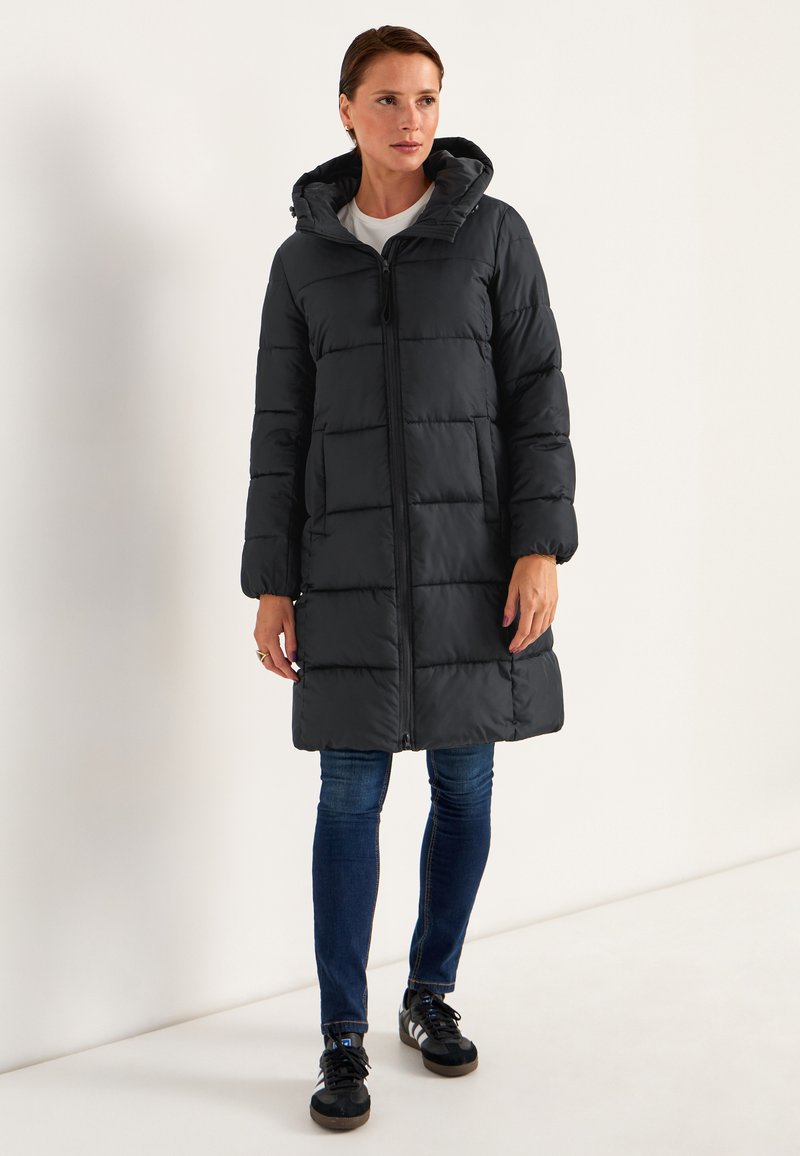 Svart puffjacka med huva, långa ärmar, dragkedja fram och horisontell quiltning. Matchad med blå jeans och svarta sneakers.