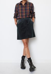 Camicia a quadri nei colori blu scuro, bordeaux e senape; gonna in velluto a coste verde scuro; abbinata a stivali neri alla caviglia e calze alte fino al ginocchio.