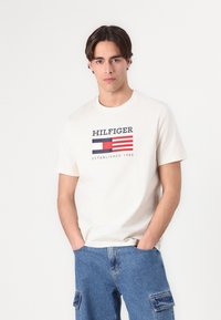 Bílá bavlněná tričko s námořnickou a červenou logem se pruhy a textem "HILFIGER ESTABLISHED 1985," doplněné světle modrými cargo šortkami.