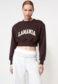 Moteris dėvi tamsią trumpą sportinę palaidinę su užrašu "LAMANIA" ir šviesiai pilkas kelnaites su raišteliais, pozuoja prieš baltą fono.
