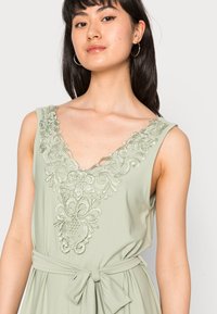 Vestido sem mangas de um verde-claro com bordados intrincados na gola e uma cintura ajustada com um laço de tecido.