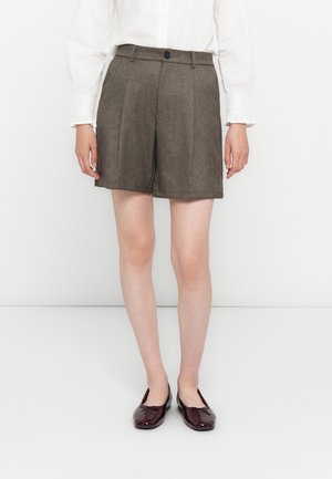 Donna che indossa pantaloncini marroni su misura, camicetta bianca abbottonata e mocassini in vernice bordeaux, in piedi davanti a uno sfondo bianco.