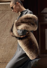 Donna con abito grigio e occhiali da sole avvolge una grande stola di pelliccia marrone attorno alle spalle, in piedi contro un muro di pietra strutturato e un portone scuro.