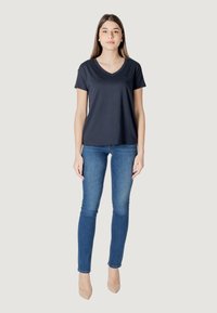 T-shirt navy a V con tessuto morbido e maniche corte, abbinato a jeans blu aderenti e tacchi alti beige, in piedi contro uno sfondo neutro.