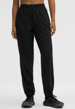 Donna che indossa pantaloni neri a vita elastica, dalla vestibilità ampia con polsini elasticizzati e scarpe da ginnastica nere su uno sfondo chiaro uniforme.