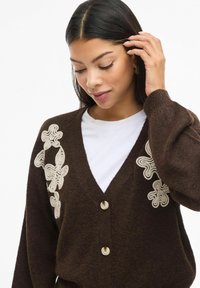 Sieviete pieskaras saviem matiem, ģērbusies tumši brūnā kardiganā ar bēšiem ziedu rakstiem virs balta krekla, skatoties uz leju ar nelielu smaidu.