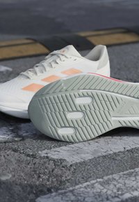 Chaussures de sport blanches et pêche sur l'asphalte, mettant en avant la semelle grise texturée avec trois découpes ovales et le logo "ADIWEAR".