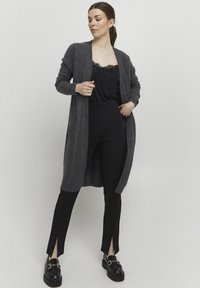 Grå lång kappa i mjukt stickat material, bärs över en svart camisole med spetsdetaljer och svarta byxor med slitsar. Svarta loafers fulländar looken.