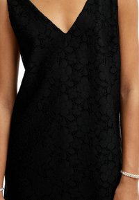 Čierna čipková šaty s V-výstrihom, botanickými vzormi, jemnou textúrou a svetlým leskom. Na zápästí je viditeľný náramok.