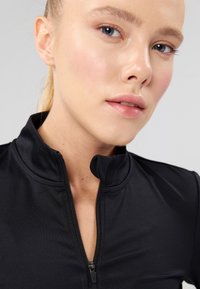 Čierny top na zips s vysokým golierom, vyrobený z hladkej, elastickej látky. Má krátky zips a priliehavý strih.