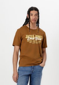 Camiseta de manga corta marrón con "JACK & JONES Denim Goods" impreso en color crema y dorado. Combinada con jeans de mezclilla azul. Textura suave.