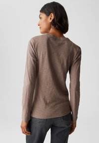 Donna con capelli corti scuri che indossa un top marrone a maniche lunghe e jeans scuri, di spalle contro uno sfondo chiaro e semplice.