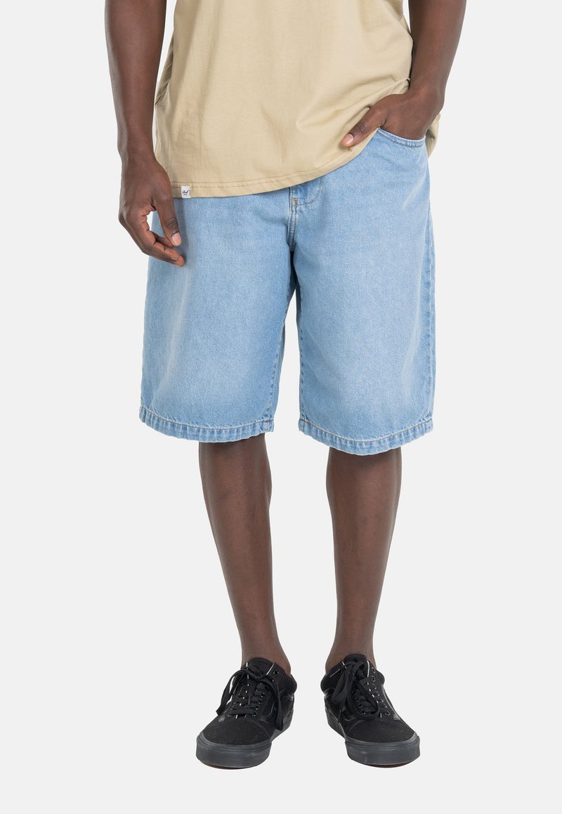 Pantalones cortos de mezclilla azul claro con un ajuste holgado, dobladillos enrollados y bolsillos laterales. Combinados con zapatillas negras y una camiseta color caqui.