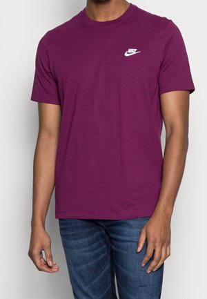 Mężczyzna ubrany w gładką fioletową koszulkę Nike z białym logo na piersi, w niebieskich dżinsach, na jednolitym jasnym tle.