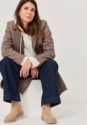 Beige suède enkellaarsjes met een ritssluiting aan de zijkant, gecombineerd met een bruine houndstooth jas en een witte hoodie, gedragen met donkerblauwe wijde jeans.