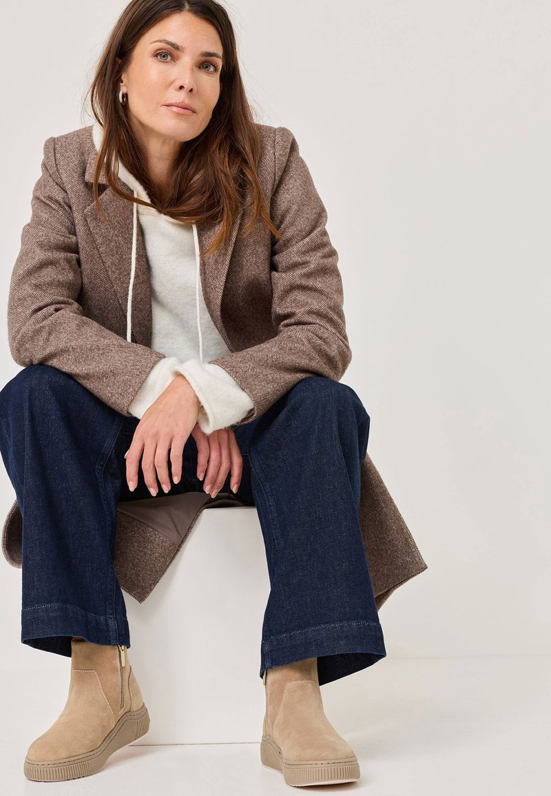 Bottines en suède beige avec une fermeture éclair sur le côté, associées à un manteau en pied-de-poule marron et un sweat à capuche blanc, portées avec un jean large bleu foncé.